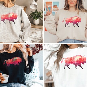May include: Four different color sweatshirts featuring a silhouette of a bison with a sunset landscape inside the silhouette. The sweatshirts are available in grey, beige, black, and white.