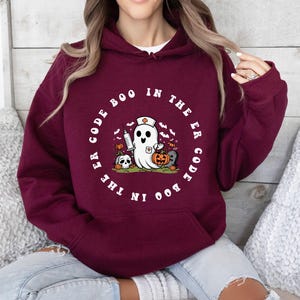 May include: A maroon hoodie with a white graphic of a ghost wearing a nurse's cap and holding a syringe. The text around the ghost reads "Boo in the ER Code the Boo in the ER Code".