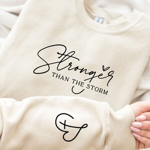 Pu&ograve; includere: Una felpa color crema con il testo "Stronger than the storm" in corsivo nero e un piccolo simbolo di cuore. La felpa ha anche un piccolo simbolo di croce sulla manica.