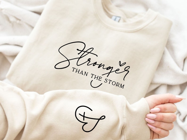 Pu&ograve; includere: Una felpa color crema con il testo "Stronger than the storm" in corsivo nero e un piccolo simbolo di cuore. La felpa ha anche un piccolo simbolo di croce sulla manica.