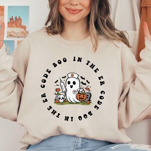 May include: A cream-colored sweatshirt with a black graphic print of a ghost wearing a nurse's hat and holding a syringe. The text around the ghost reads "Code Boo in the ER Code Boo in the ER".