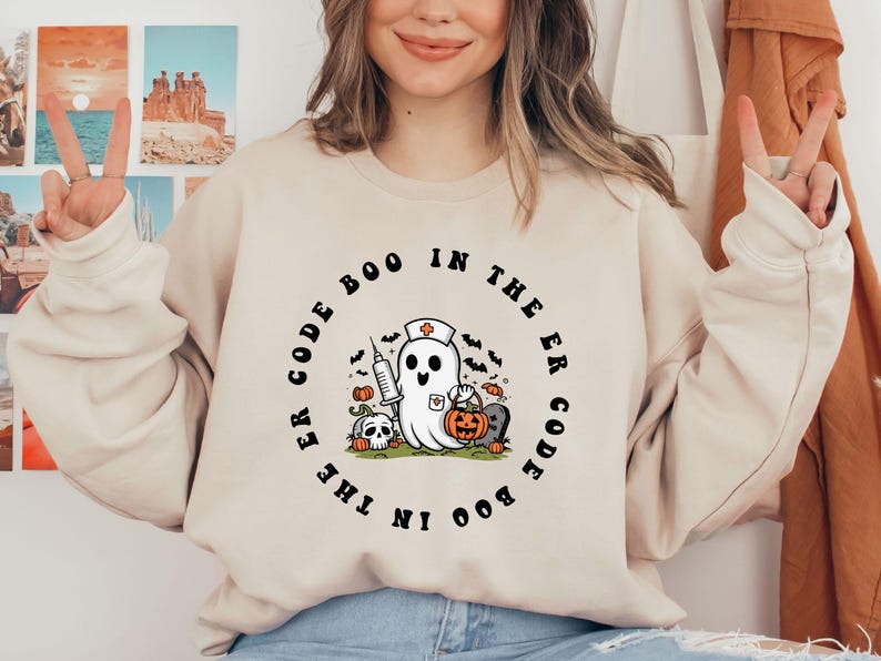 May include: A cream-colored sweatshirt with a black graphic print of a ghost wearing a nurse's hat and holding a syringe. The text around the ghost reads "Code Boo in the ER Code Boo in the ER".
