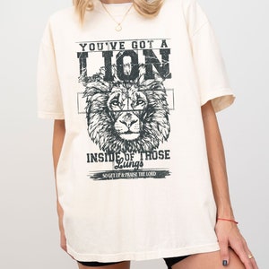 May include: A cream colored t-shirt with a black and white graphic of a lion's head. The text "YOU'VE GOT A LION INSIDE OF THOSE Lungs SO GET UP & PRAISE THE LORD" is printed on the shirt.