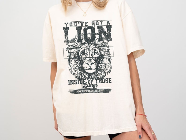 May include: A cream colored t-shirt with a black and white graphic of a lion's head. The text "YOU'VE GOT A LION INSIDE OF THOSE Lungs SO GET UP & PRAISE THE LORD" is printed on the shirt.