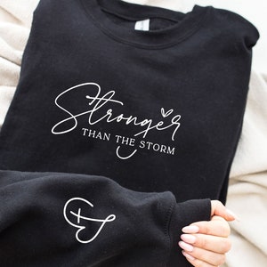 Pu&ograve; includere: Felpa nera con testo bianco che recita "Stronger than the storm" e un piccolo cuore. La manica ha un design a croce e cuore bianco.