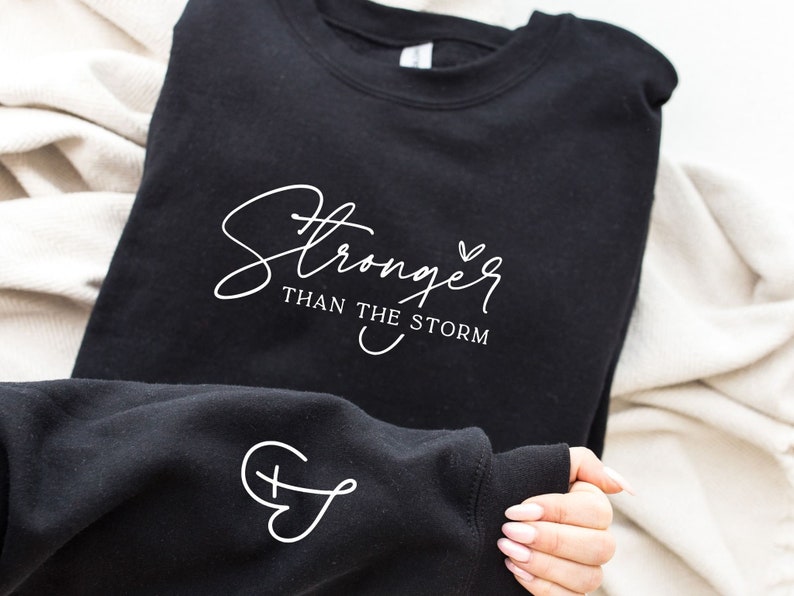 Pu&ograve; includere: Felpa nera con testo bianco che recita "Stronger than the storm" e un piccolo cuore. La manica ha un design a croce e cuore bianco.