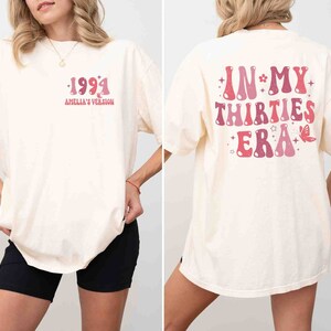 May include: A white oversized t-shirt with a pink graphic on the front that says "1994 Amelia's Version" and a pink graphic on the back that says "In My Thirties Era" with a butterfly and stars.