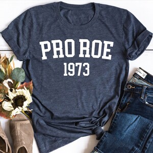 May include: A navy blue t-shirt with white text that reads "PRO ROE 1973". The shirt is laid flat on a white wooden surface with a pair of brown sandals and blue jeans.