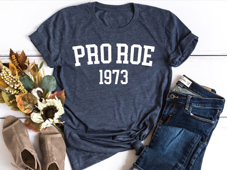 May include: A navy blue t-shirt with white text that reads "PRO ROE 1973". The shirt is laid flat on a white wooden surface with a pair of brown sandals and blue jeans.