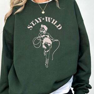 May include: A dark green sweatshirt with a pink graphic of a woman in a cowboy hat and the text "STAY WILD" printed on the front.