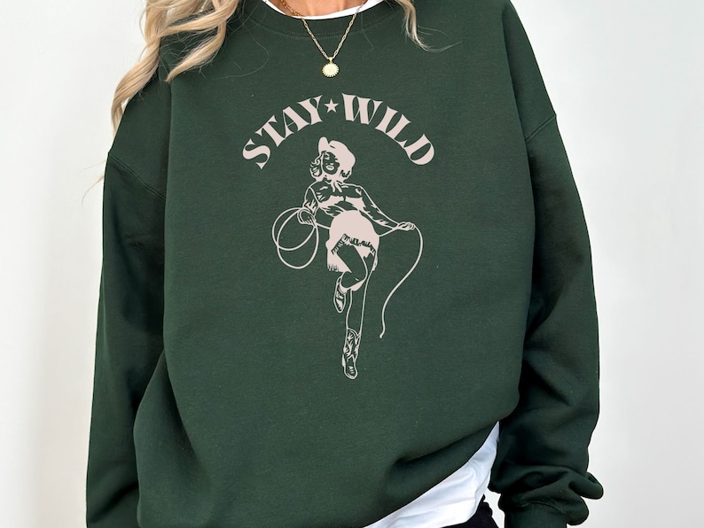 May include: A dark green sweatshirt with a pink graphic of a woman in a cowboy hat and the text "STAY WILD" printed on the front.