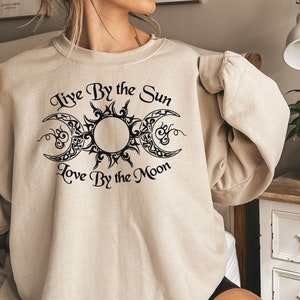 May include: Beige sweatshirt with a black sun and moon graphic. The text "Live By the Sun, Love By the Moon" is printed above and below the graphic. The sweatshirt is a neutral color and has long sleeves.