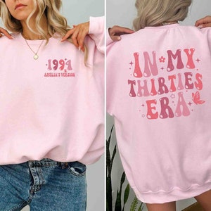 May include: Pink sweatshirt with a retro-style graphic on the front that says "1991 Amelia's Version" and a retro-style graphic on the back that says "In My Thirties Era".