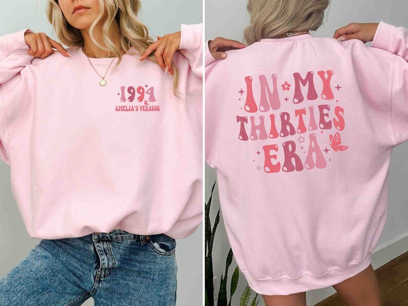 May include: Pink sweatshirt with a retro-style graphic on the front that says "1991 Amelia's Version" and a retro-style graphic on the back that says "In My Thirties Era".