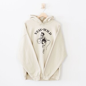 May include: A cream-colored hoodie with a black graphic of a woman in a cowboy hat holding a lasso. The graphic says "Stay Wild".