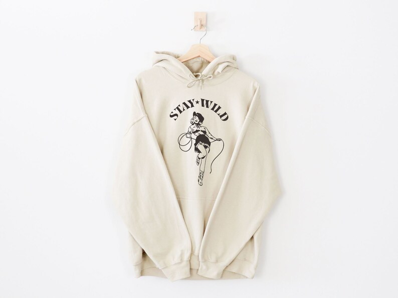 May include: A cream-colored hoodie with a black graphic of a woman in a cowboy hat holding a lasso. The graphic says "Stay Wild".