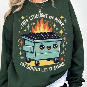May include: A dark green sweatshirt with a cartoon image of a green dumpster with a smiling face and flames coming out of the top. The text "This little light of mine, I'm gonna let it shine" is printed on the sweatshirt.