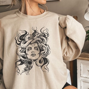 May include: A beige sweatshirt with a black and white graphic print of Medusa, a mythical Greek figure with snakes for hair.