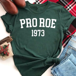 May include: A dark green t-shirt with white text that reads "PRO ROE 1973". The shirt is laid out on a white surface with a plaid blanket, a pair of brown shoes, and a small gift box with a red and white plaid bow.