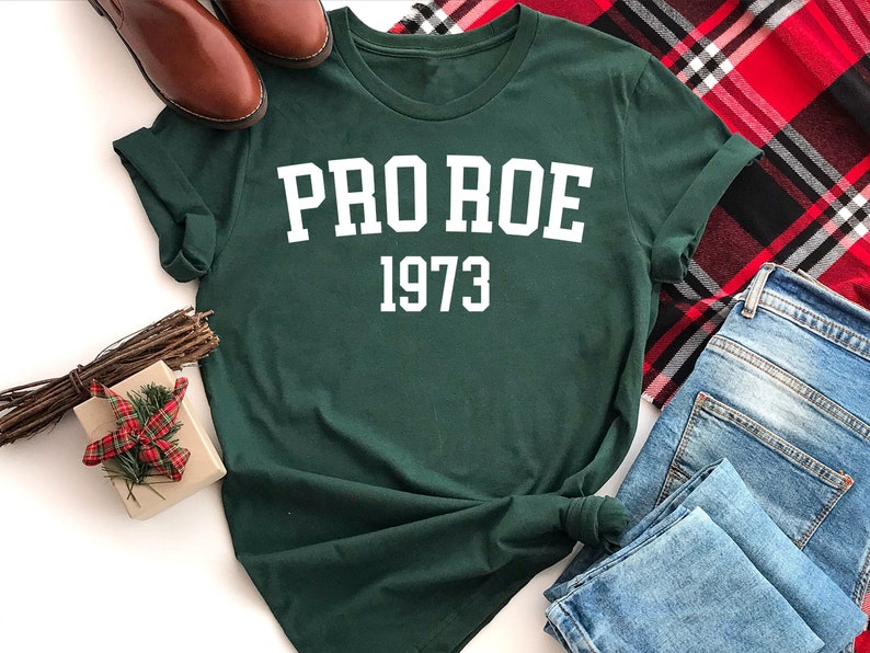 May include: A dark green t-shirt with white text that reads "PRO ROE 1973". The shirt is laid out on a white surface with a plaid blanket, a pair of brown shoes, and a small gift box with a red and white plaid bow.
