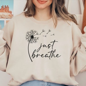 May include: A beige sweatshirt with the text "just breathe" and a dandelion graphic. The sleeves have the words "inhale" and "exhale" printed on them.