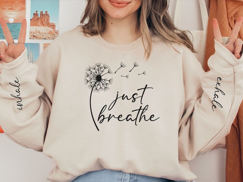 May include: A beige sweatshirt with the text "just breathe" and a dandelion graphic. The sleeves have the words "inhale" and "exhale" printed on them.