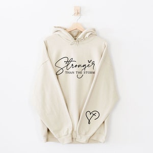 Pu&ograve; includere: Una felpa con cappuccio beige con il testo "Stronger than the storm" in corsivo nero e un piccolo cuore con una croce all'interno sul braccio sinistro.
