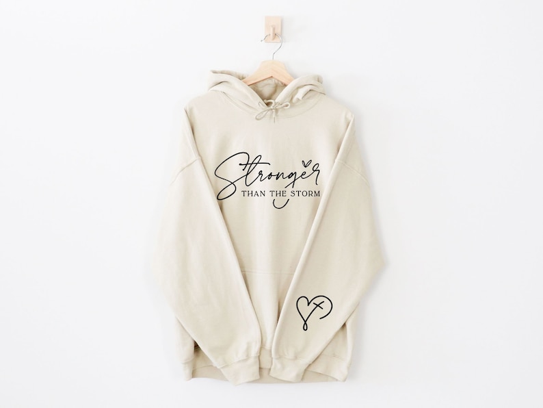 Pu&ograve; includere: Una felpa con cappuccio beige con il testo "Stronger than the storm" in corsivo nero e un piccolo cuore con una croce all'interno sul braccio sinistro.