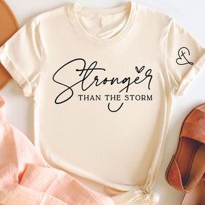 Pu&ograve; includere: Una maglietta color crema con il testo "Stronger than the storm" in corsivo nero. Un piccolo disegno a forma di cuore e croce si trova sulla manica destra.