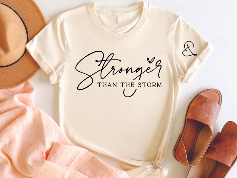 Pu&ograve; includere: Una maglietta color crema con il testo "Stronger than the storm" in corsivo nero. Un piccolo disegno a forma di cuore e croce si trova sulla manica destra.