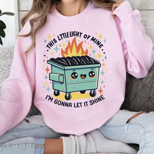 May include: Pink sweatshirt with a graphic of a cartoon dumpster with a smiling face and flames. The text on the sweatshirt reads "This little light of mine... I'm gonna let it shine."