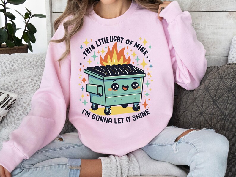 May include: Pink sweatshirt with a graphic of a cartoon dumpster with a smiling face and flames. The text on the sweatshirt reads "This little light of mine... I'm gonna let it shine."