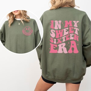 May include: Green sweatshirt with a pink smiley face on the front and the text "In My Sweet Sixteen Era" in pink retro font on the back.