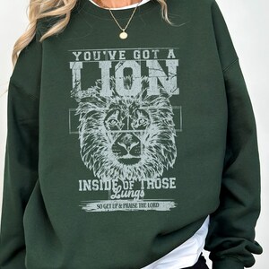 May include: A dark green sweatshirt with a white graphic print of a lion's head and the text "You've got a lion inside of those lungs. So get up & praise the Lord."
