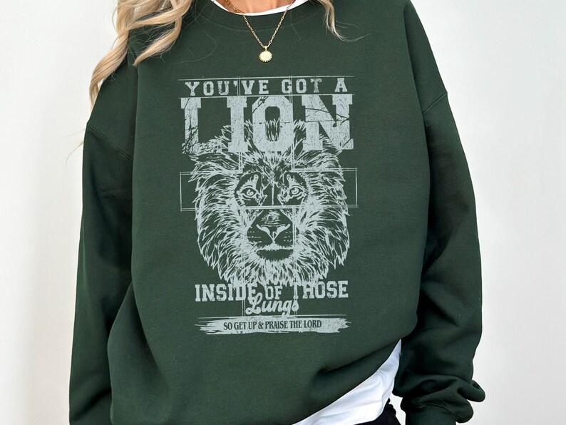May include: A dark green sweatshirt with a white graphic print of a lion's head and the text "You've got a lion inside of those lungs. So get up & praise the Lord."
