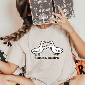 May include: A woman wearing a white t-shirt with a graphic of two geese facing each other with the text "Goose Bumps". She is holding a book in front of her face. The book is black with white text that reads "Spells & Potions".