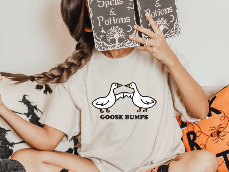 May include: A woman wearing a white t-shirt with a graphic of two geese facing each other with the text "Goose Bumps". She is holding a book in front of her face. The book is black with white text that reads "Spells & Potions".