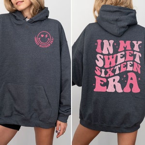 May include: A dark gray hoodie with a pink smiley face and the text "Sweet Sixteen Era" on the front and the text "In My Sweet Sixteen Era" on the back.