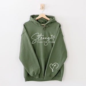 Pu&ograve; includere: Una felpa con cappuccio verde con il testo "Stronger than the storm" e un cuore con una croce all'interno sulla manica.