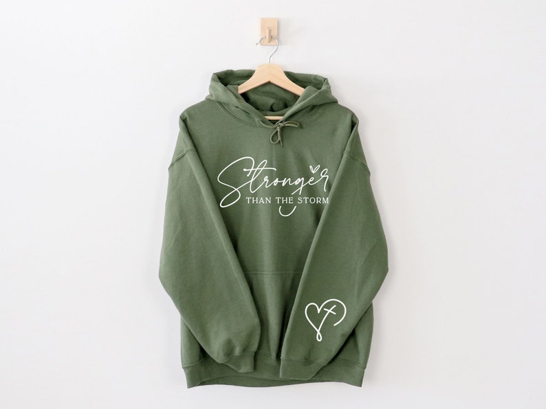 Pu&ograve; includere: Una felpa con cappuccio verde con il testo "Stronger than the storm" e un cuore con una croce all'interno sulla manica.