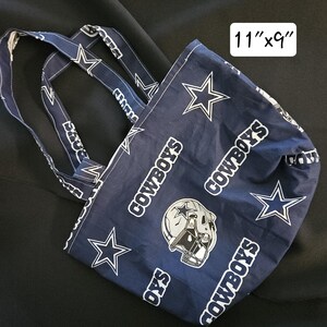 Pu&ograve; includere: Una borsa di tela blu e bianca con il logo dei Dallas Cowboys e le parole "Cowboys" ripetute sulla borsa. La borsa misura 28 cm per 23 cm.