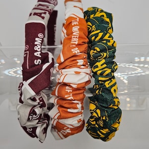 May include: Three fabric hair scrunchies with different college logos. The scrunchies are maroon and white, orange and white, and green and yellow.