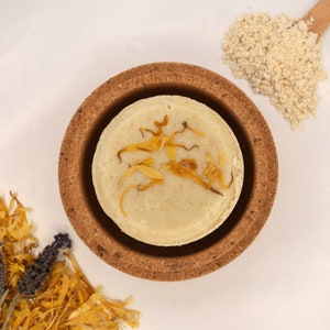 May include: A round, light-colored shampoo bar with orange flower petals, resting in a brown cork dish. A pile of beige powder and dried orange petals are in the background, all on a white surface. The shampoo bar is a natural beauty product.