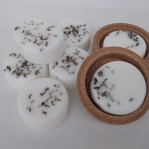 以下が含まれることがあります： 乾燥ハーブを散らした白い丸いワックスメルトのクローズアップ。コルクコースターに入っているものもあります。ワックスメルトは白い表面に配置されています。画像は明るく、ワックスとハーブの質感がわかります。