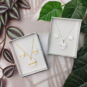 Könnte beinhalten: Zwei Schmucksets in silbernen Boxen. Jedes Set enthält eine Halskette und Ohrringe mit weißen Keramikanhängern und goldenen Akzenten.