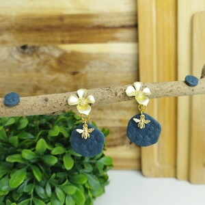 Könnte beinhalten: Ein Paar blaue und goldene Ohrringe mit einem Blumen- und einem Bienenanhänger. Die Ohrringe hängen an einem Holzast.