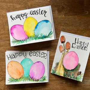 May include: Three handmade Easter cards with watercolor illustrations. The first card features three pastel colored eggs in a nest of green grass. The second card features three pastel colored eggs in a nest of green grass. The third card features a single pink egg with brown mushrooms growing from a tree stump.