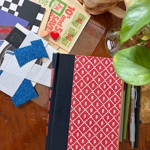 May include: A hardcover book with a black spine and a red cover featuring a white diamond lattice pattern with small white flowers. Beside the book are various paper scraps, including blue glitter rectangles, grid paper, and vintage S&H Green Stamps.
