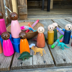 May include: A set of wooden peg dolls with different facial expressions, including happy, sad, angry, and surprised. The dolls are painted in various colors, including pink, orange, red, blue, and green. There are also small plastic toy dinosaurs and a lion on the wooden surface.