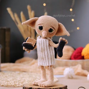 May include: A handmade crochet doll of a character with large ears, green eyes, and a sad expression. The doll wears a white tunic and holds a black sock and a brown object. It stands on a wooden base.